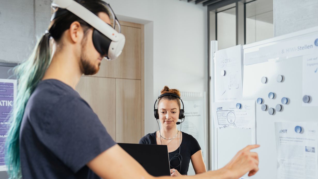 Amadeus Bramsiepe, KIT Person mit VR-Brille zeigt auf ein Schwarzes Brett mit Aushängen. Im Hintergrund steht eine Person mit Headset und schaut auf ihren Laptop.